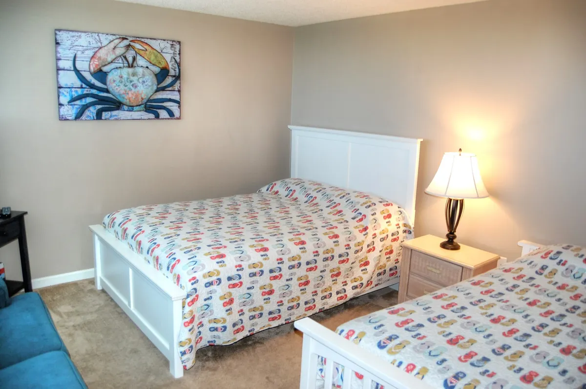 A cheerful bedroom with two beds dressed in colorful bedspreads, a bedside lamp for nighttime reading, and playful crab artwork on the wall. The space feels light-hearted and comfortable, perfect for families or children.