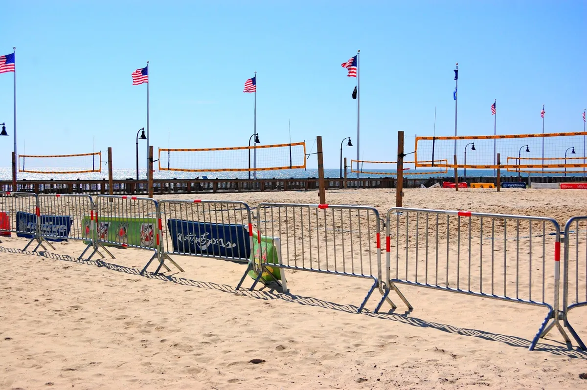 An energetic beachside scene featuring sand volleyball courts, American flags, and visitors sunbathing and playing. The ocean and wide sandy beach create a classic Myrtle Beach atmosphere.