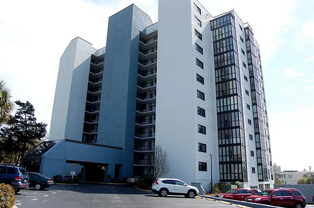 This image displays the exterior of Seamark Towers, showcasing its tall, contemporary building structure with large glass windows and an inviting parking area in the foreground. The building stands out in the mild coastal sunlight, highlighting a sense of arrival and community.