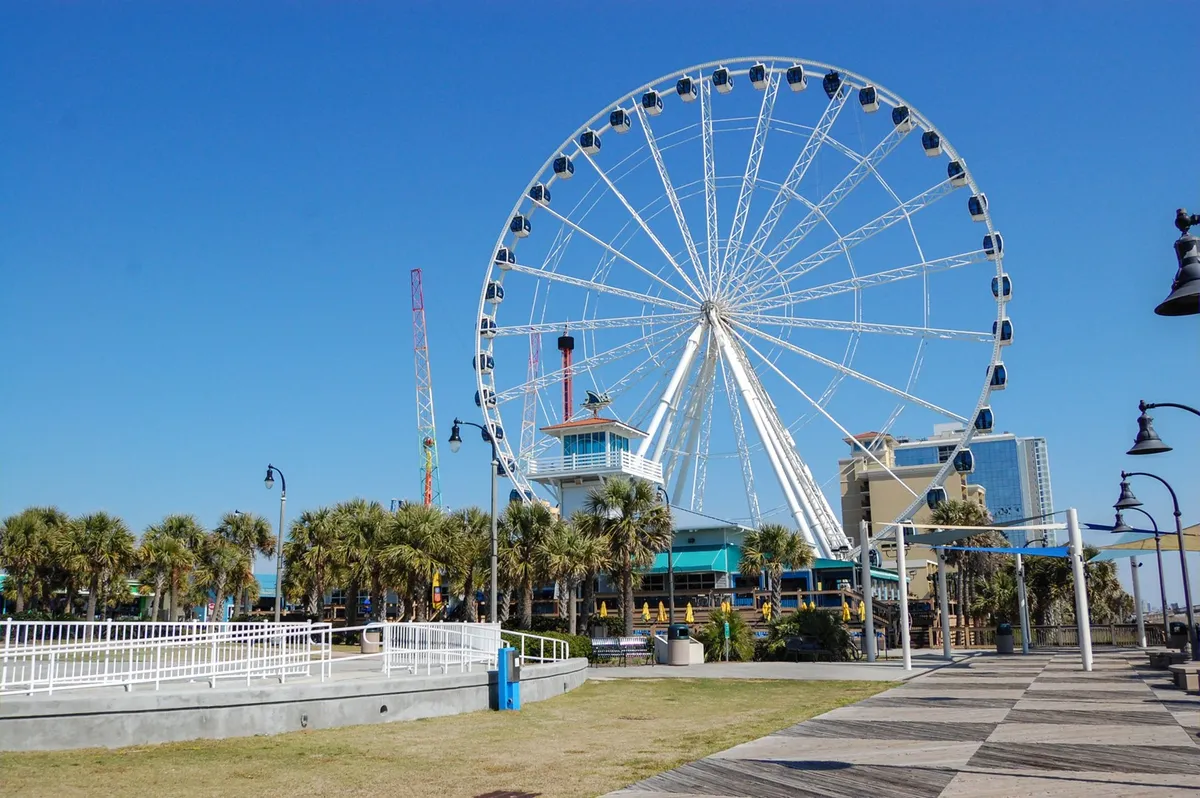 A vibrant exterior scene featuring the Myrtle Beach SkyWheel, with palm trees lining the boardwalk and an amusement park atmosphere. The Ferris wheel stands tall as a local landmark, bustling with visitors enjoying rides and the seaside excitement.