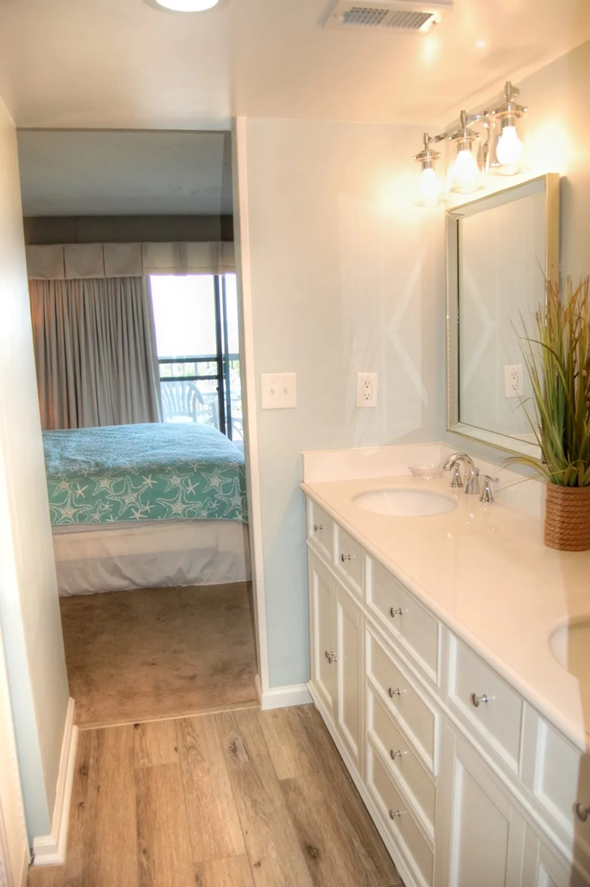 A modern bathroom with a spacious double-sink vanity, large mirror, and decorative plant. The view extends through a doorway into a calming bedroom space, blending privacy and convenience for guests.