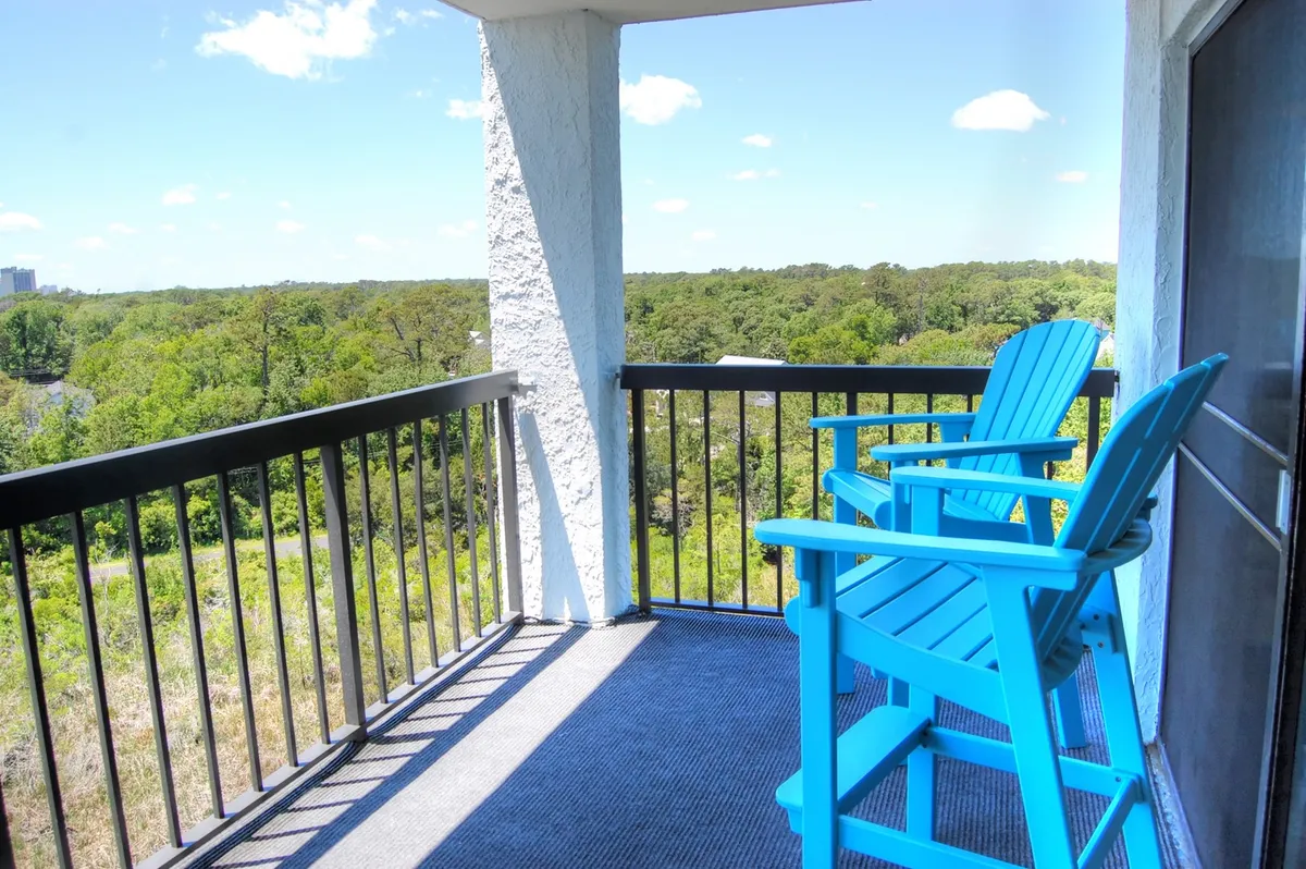 A balcony setting featuring blue Adirondack chairs overlooks a green landscape, framed by a sturdy railing. The tranquil mood suggests a comfortable spot for watching sunrises or unwinding after a day at the beach, connecting indoor relaxation with the area’s natural beauty.