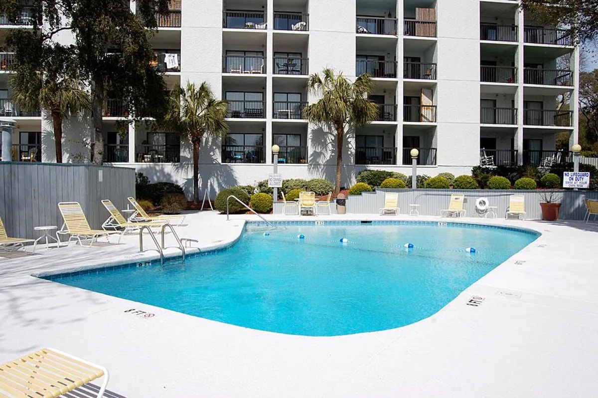 A vibrant outdoor pool surrounded by lounge chairs and palm trees, with balconies overlooking the scene. The setting exudes the inviting atmosphere of a resort, perfect for swimming, sunbathing, and socializing in a lush, tropical environment.