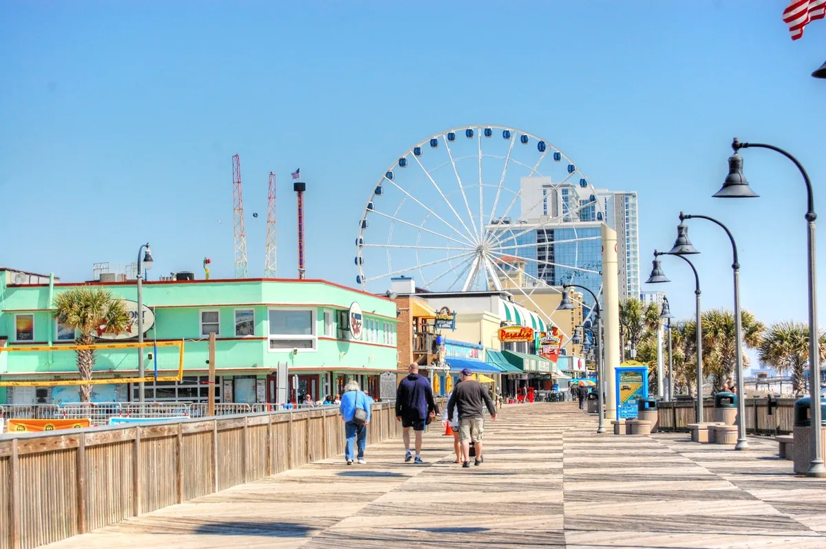 A festive, colorful exterior shot showcasing the Ferris wheel, palm trees, vibrant boardwalk shops, and people strolling and socializing. The energy of Myrtle Beach’s entertainment district is front and center.