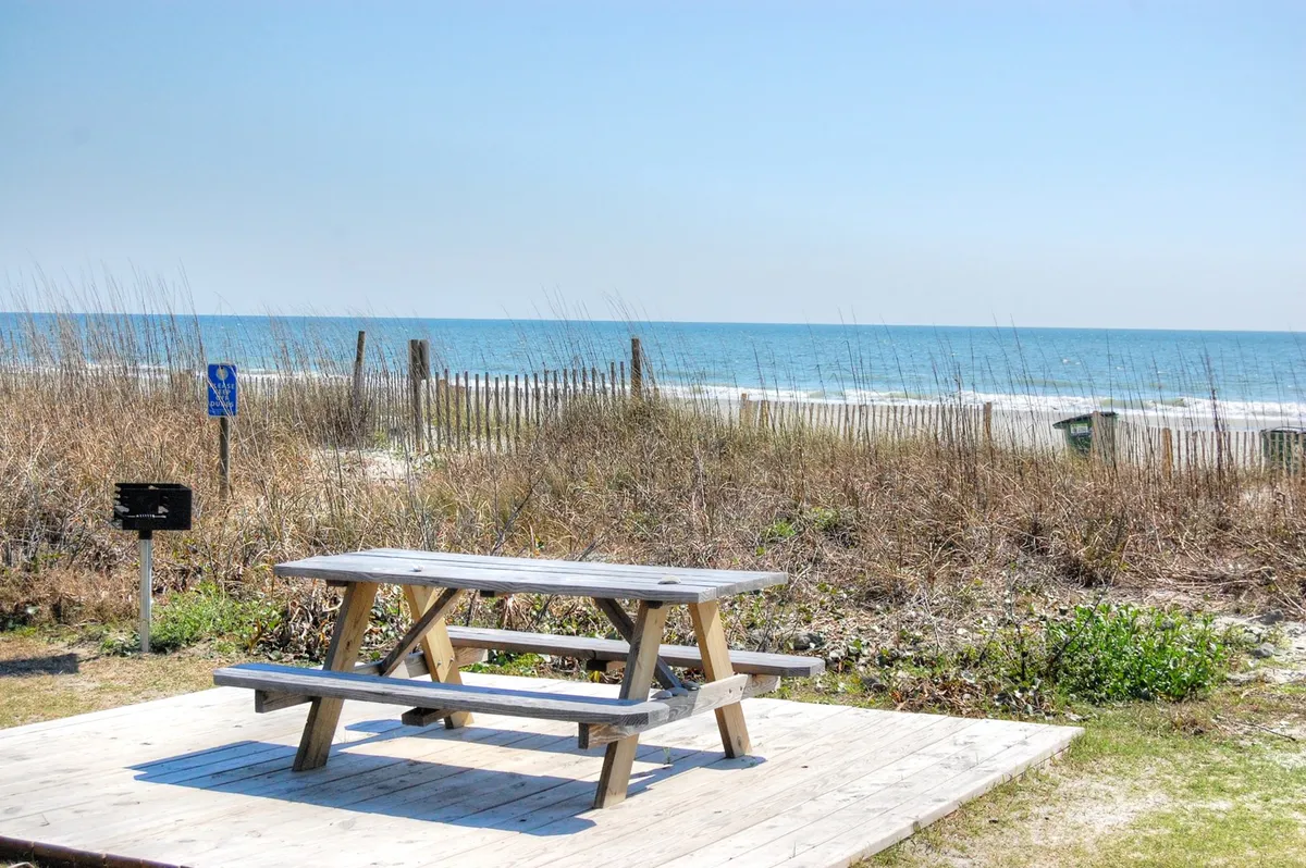 A wooden picnic table sits on the sand dunes overlooking the sparkling ocean, bordered by fencing that adds a natural, coastal feel. The backdrop features tranquil waves and wide, open skies. The table is positioned perfectly for family or group picnics, making the most of beachfront living.
