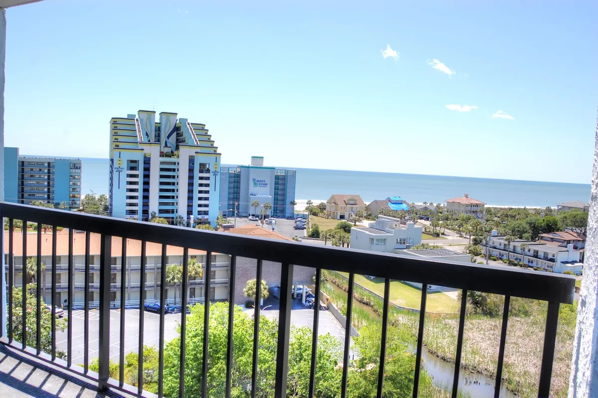 A sweeping view from a balcony reveals a panoramic scene of the ocean, the Myrtle Beach skyline, and lush greenery below. The vantage point suggests the elevated perspective of a high-rise, emphasizing the proximity to the water while highlighting the tranquil atmosphere of the Golden Mile area.