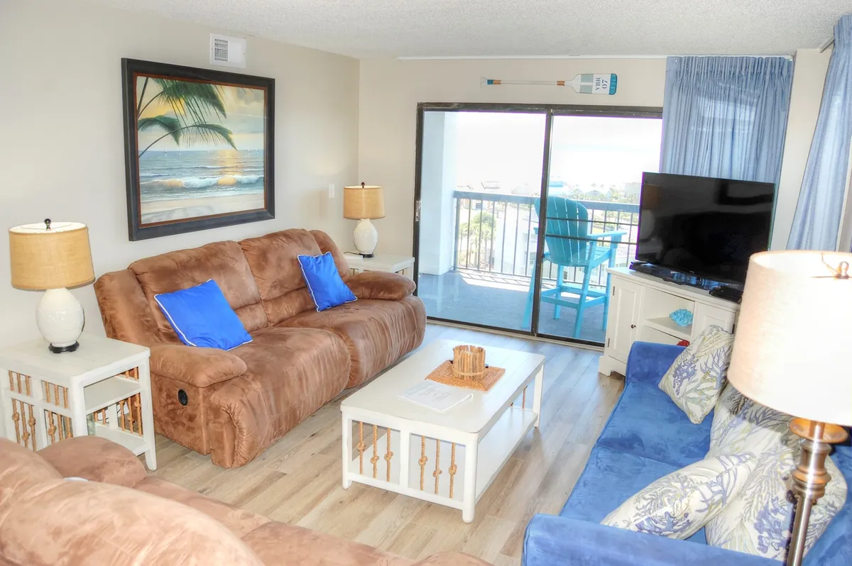 This image showcases a comfortable living room with a plush sofa, a coffee table, and a large TV. The space is bathed in natural light from expansive windows, which offer a stunning view of the ocean and access to the private balcony. The setting is perfect for relaxing, movie nights, or socializing while enjoying the beautiful scenery beyond the glass doors.