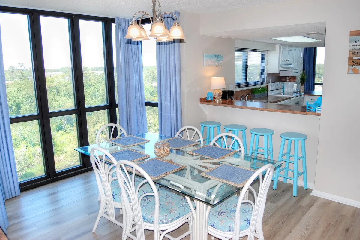 An airy dining space with a glass dining table, blue bar stools, large windows letting in abundant sunlight, and a decorative lamp. The setup is perfect for family meals and social gatherings, with comfortable seating and a relaxed, light-filled ambiance.