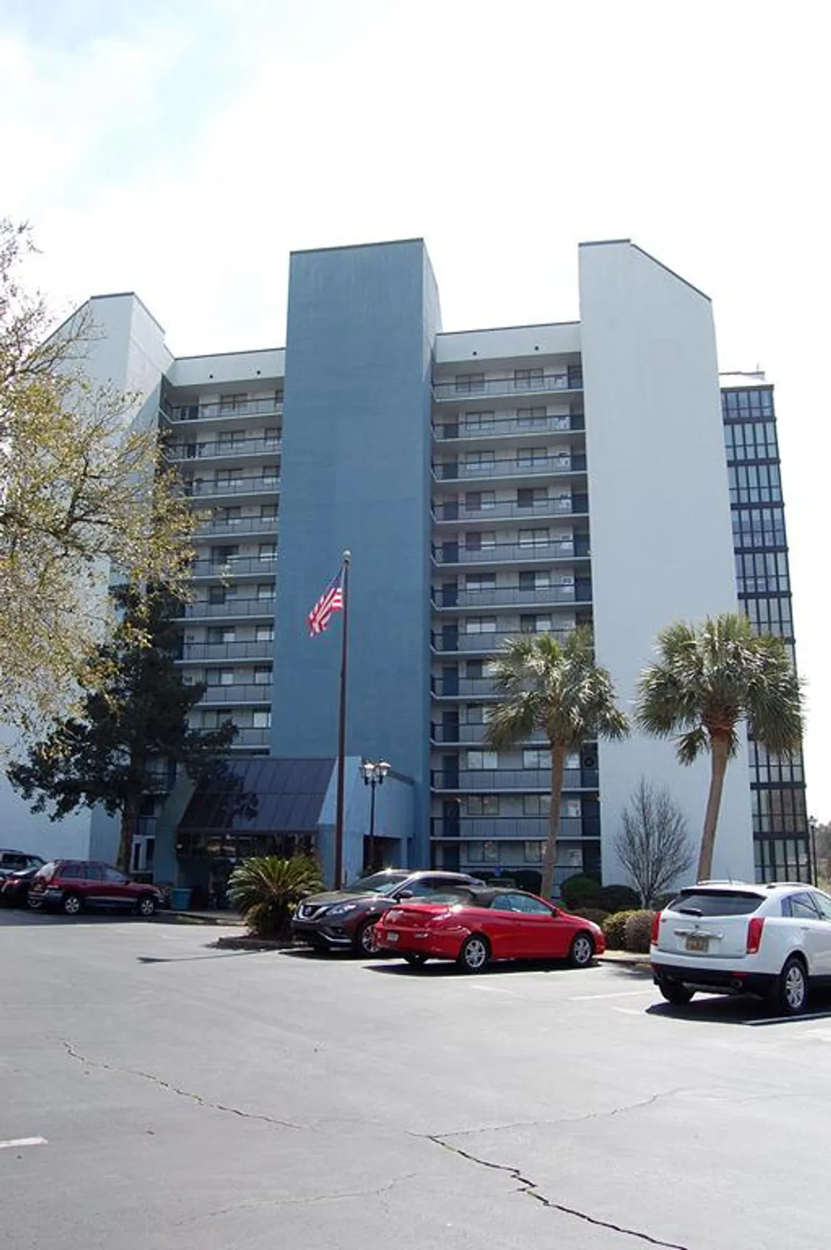 The exterior of Seamark Towers is shown with its modern tall structure, palm trees, and an inviting entrance complete with an American flag and ample parking. The sunny setting highlights the resort's vibrant atmosphere and its accessibility.