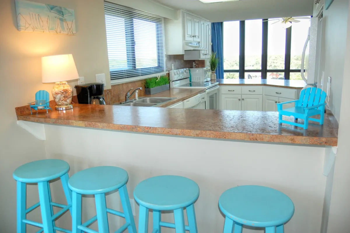 A bright kitchen area with a bar counter, blue stools, and visible appliances. The space includes a window view, adding to the cheerful and inviting atmosphere, and highlights both informal dining and socializing zones.