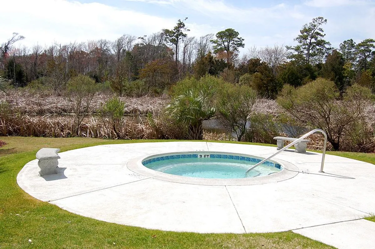 A serene hot tub nestled amidst greenery with a tranquil water view beyond. The scene invites guests to unwind and enjoy peaceful moments, whether soaking solo or chatting with friends as the sun sets.